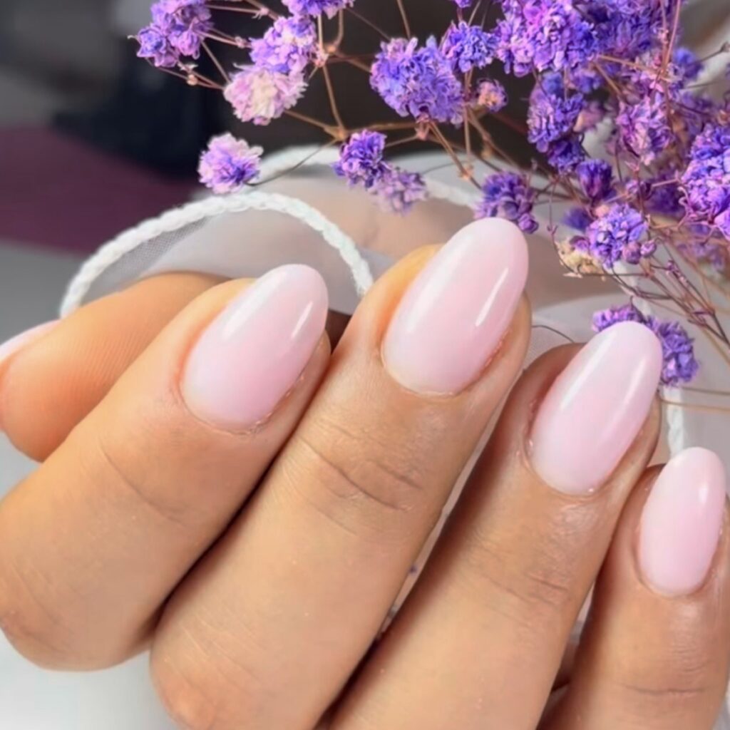 Manos con uñas almendradas en rosa bebé junto a unas flores lilas